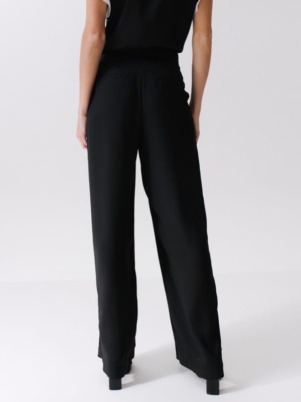 O que combina com blusa social feminina: modelo vestindo uma calça pantalona em crepe caimento fluido preta - costas.